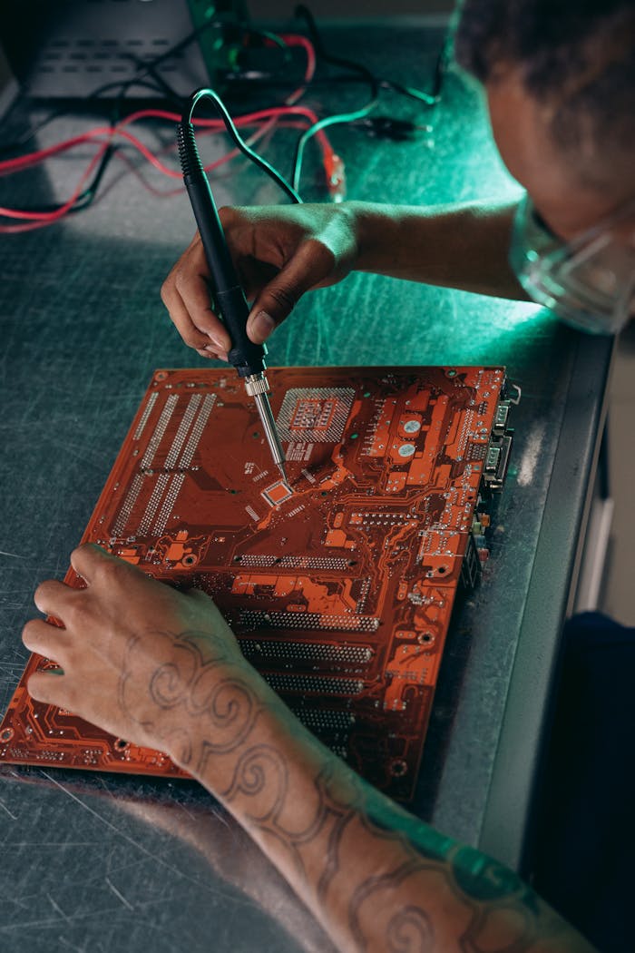 Tattooed engineer working on a motherboard with a soldering iron in a tech workshop.
